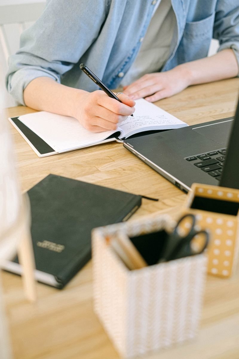 Person handwriting notes in a notebook next to a laptop while drafting an article Person handwriting notes in a notebook next to a laptop while drafting an article
